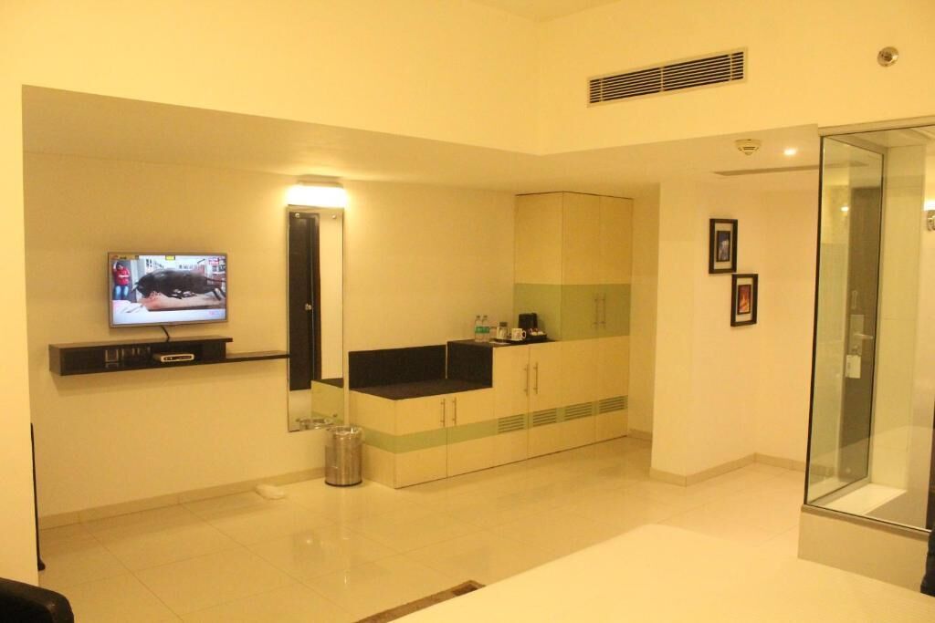A modern hotel room with a flat-screen TV mounted on the wall, a minibar, a glass-enclosed shower area, and a small trash bin.