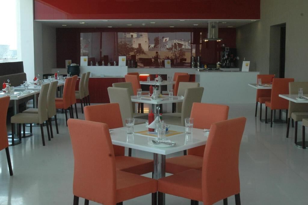 Un restaurante moderno con sillas anaranjadas y mesas blancas, un área de bar al fondo, y vistas a la ciudad a través de amplios ventanales.