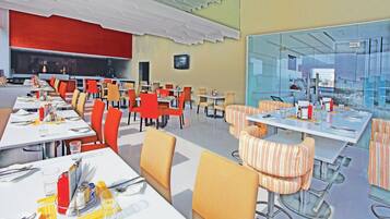 Un ristorante moderno con diverse aree dedicate alla ristorazione, tra cui un lungo tavolo condiviso e diversi tavoli più piccoli con sedute imbottite.
