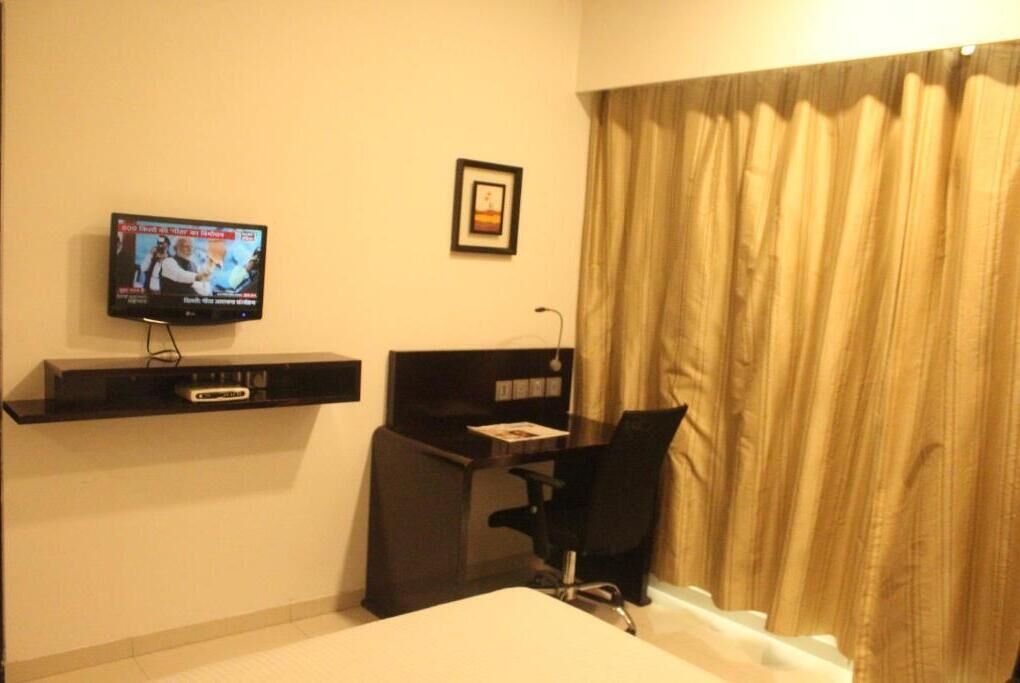 Quarto de hotel com cama, escrivaninha com cadeira, televisão fixada na parede e janela com cortinas.