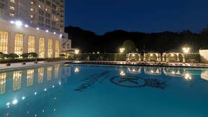 Una vista nocturna de una piscina de hotel con reflejo cristalino, rodeada de edificios bien iluminados y un paisaje montañoso.