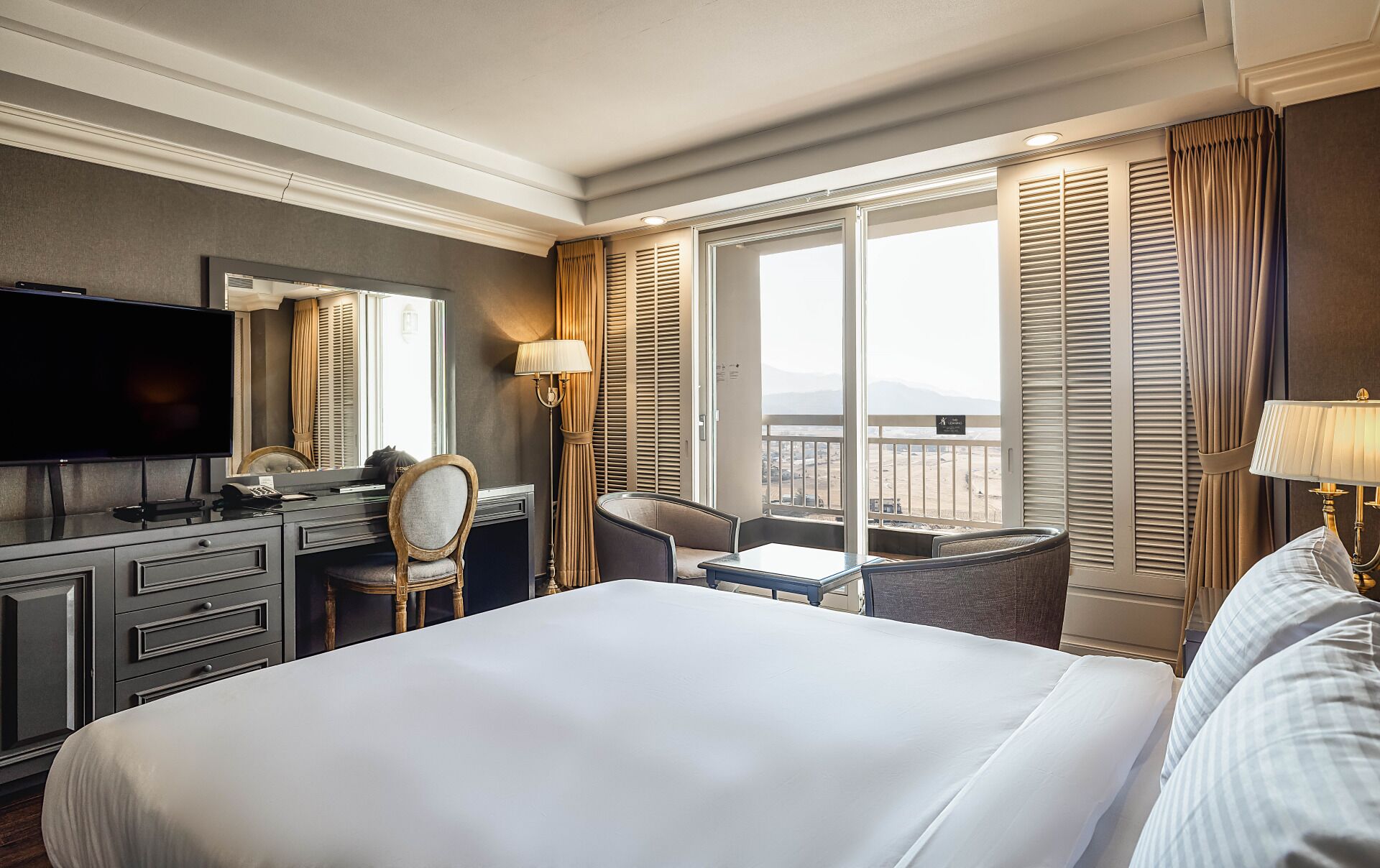 A hotel room with a large bed, a desk with a chair, a television, and a view of the outdoors.