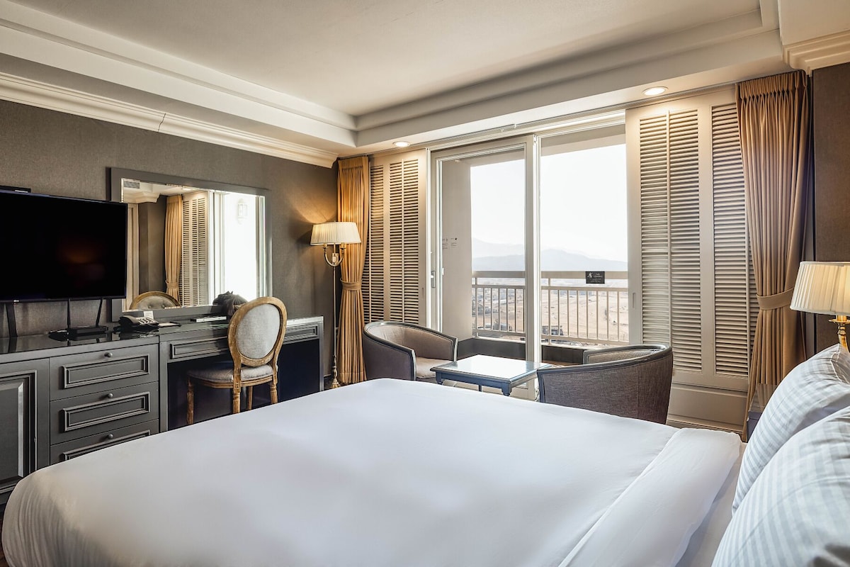 A hotel room with a large bed, a desk with a chair, a television, and a balcony with a view.