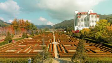 Un jardín formal con setos geométricos, un sendero central y una vista distante de un edificio moderno con tejado rojo.