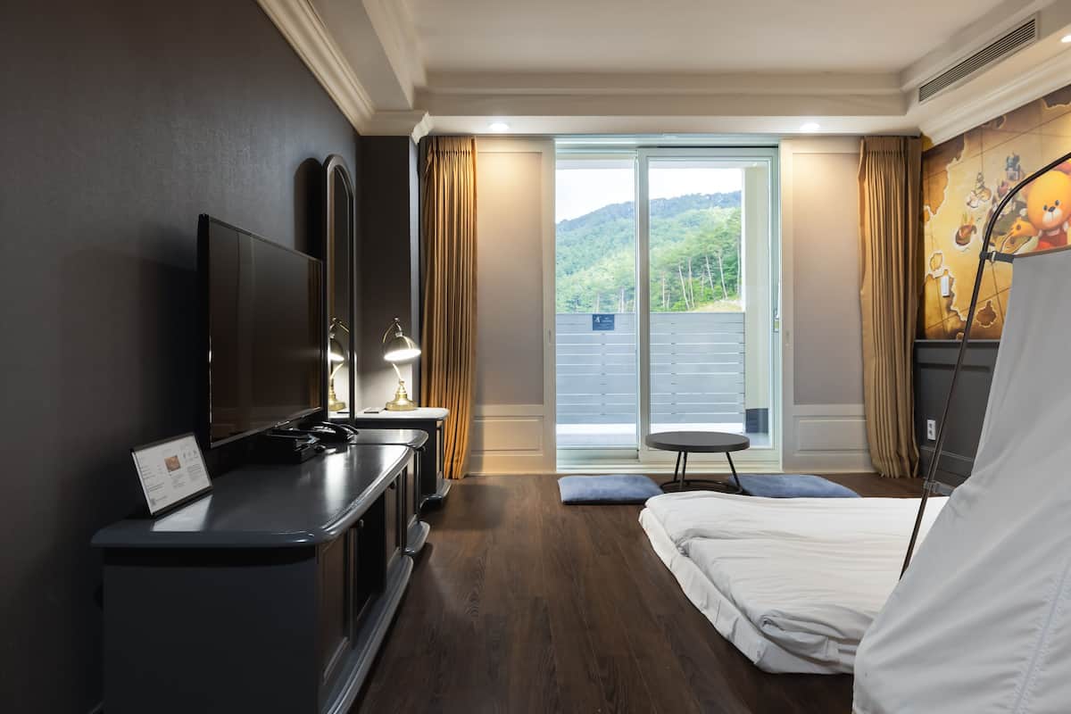 A hotel room with a bed, a TV, a desk, and a view of a mountainous landscape through the window.
