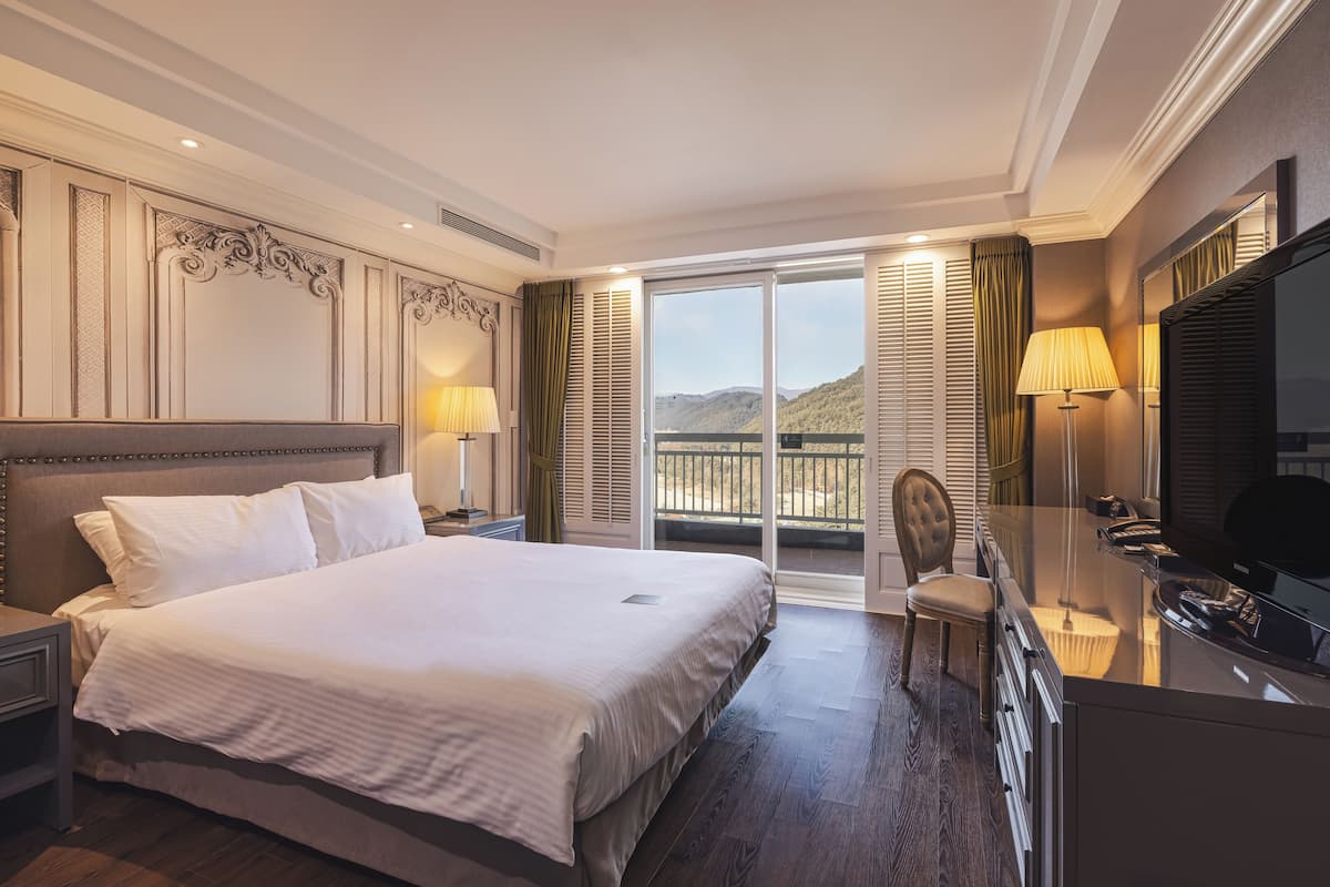 A hotel room with a large bed, a desk with a TV, a chair, and a balcony with a view of the mountains.