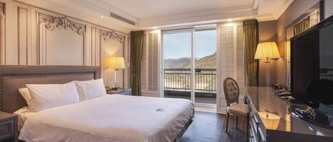 Habitación de hotel con una cama grande, un escritorio con televisor, una silla y balcón con vistas a las montañas.