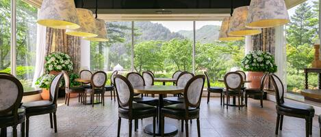 A dining area with round tables and chairs, large hanging lamps, and a view of greenery through large windows.