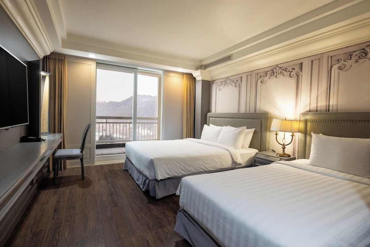 A hotel room with two beds, a desk, a chair, a TV, a balcony with a view, and a decorative headboard.