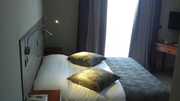 Ett hotellrum med en säng, en sänglampa, ett nattduksbord, ett skrivbord med en lampa och ett fönster med gardiner.