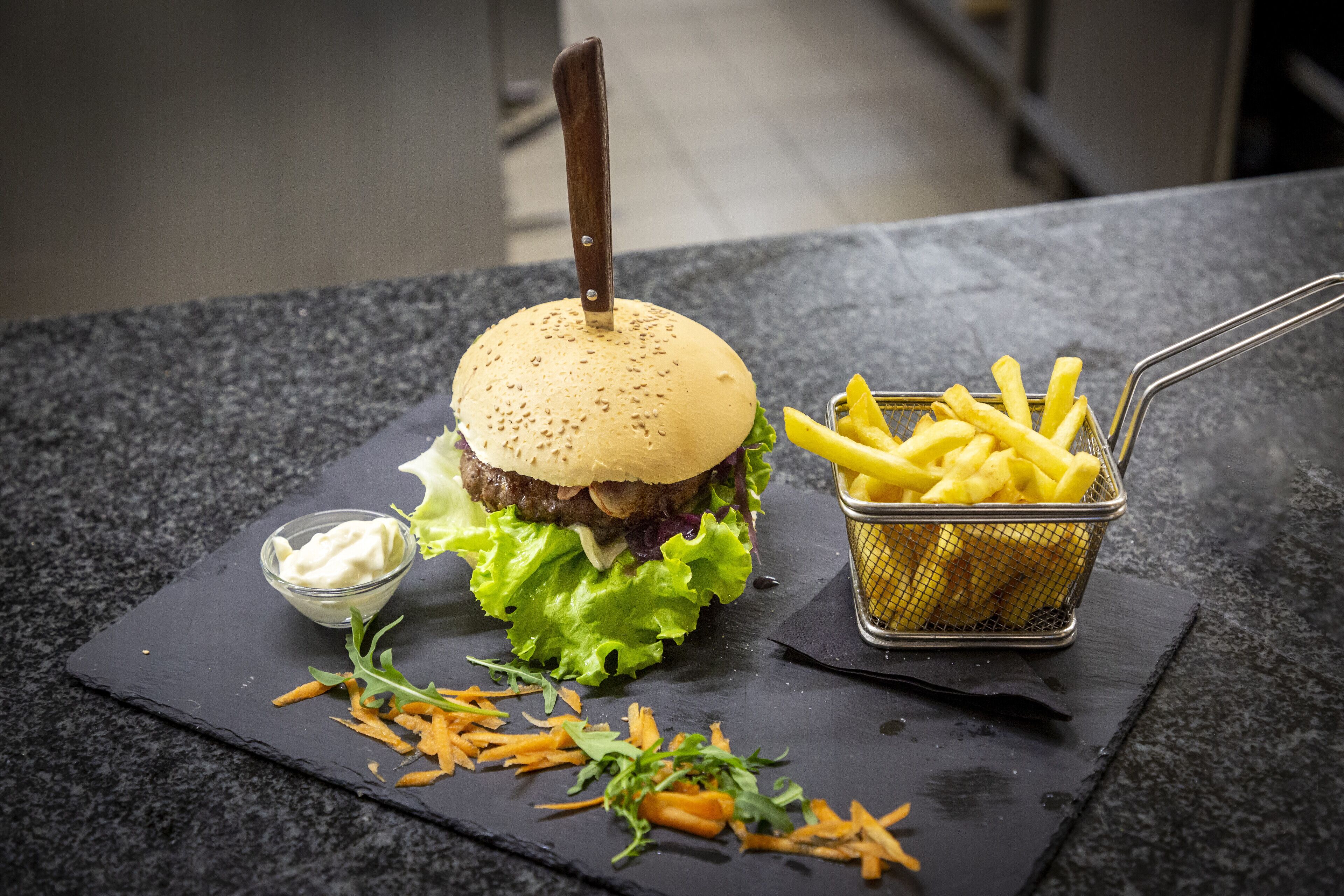 A burger with a knife on top, fries in a basket, and a small bowl of sauce on a slate serving board.