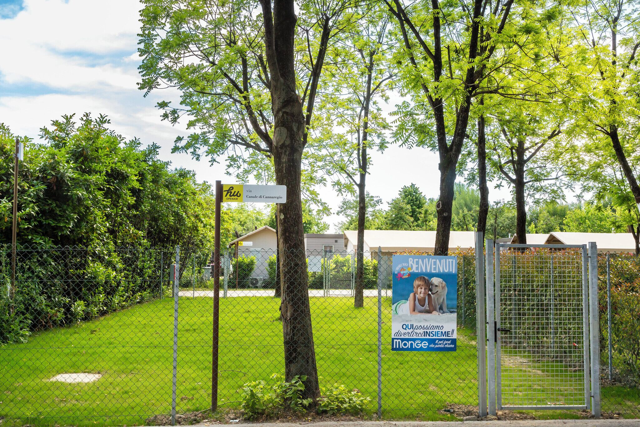 Ein von einer Hecke umgebenes Außenareal mit Bäumen, einem Schild für 'Via Canale del Cannaregio' und einem Plakat, das 'BENVENUTI' bewirbt.