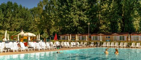 Piscina com espreguiçadeiras e guarda-sóis, cercada por árvores e um edifício com telhado vermelho.