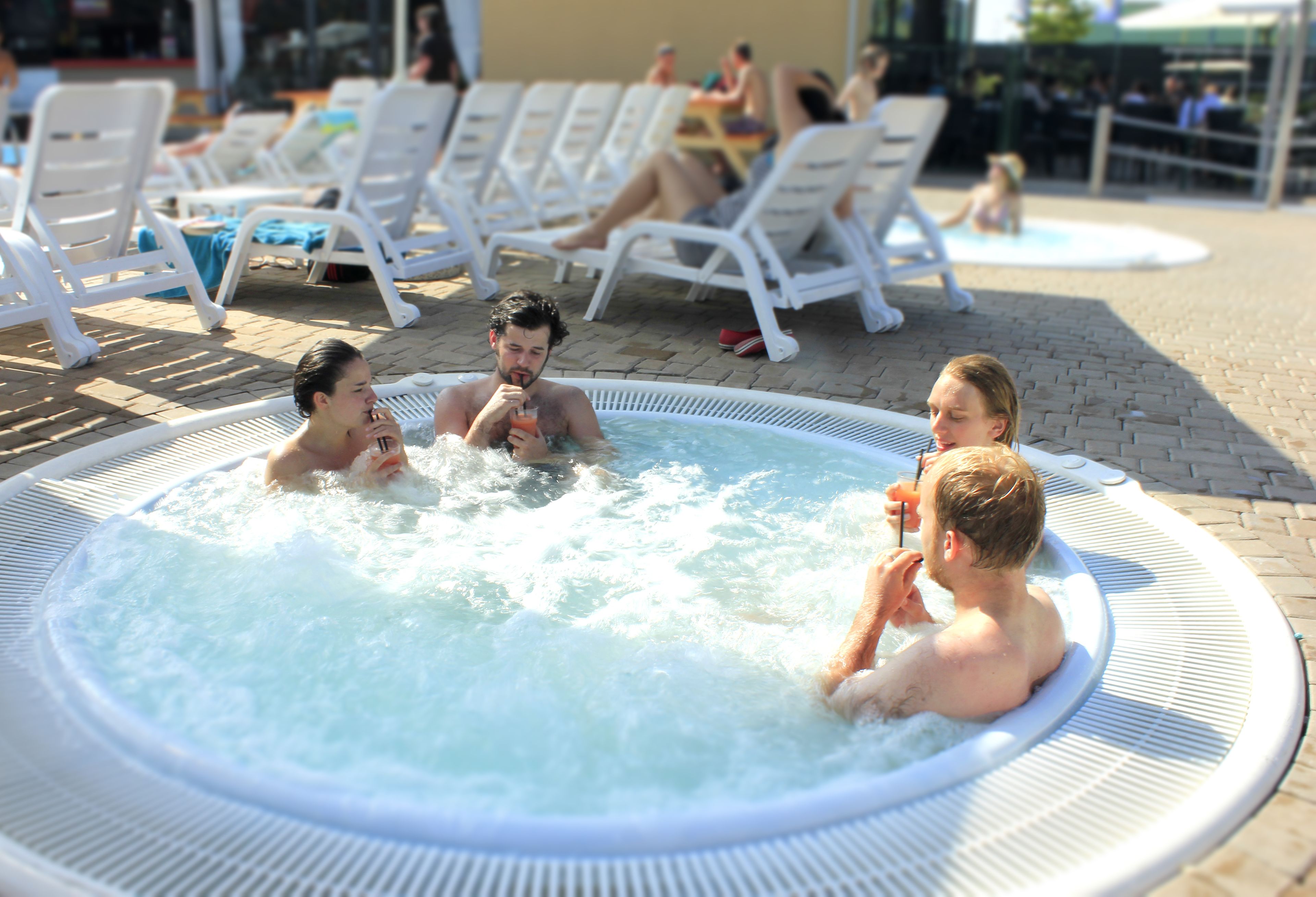 A hot tub with four people in it, surrounded by white lounge chairs and a pool area.