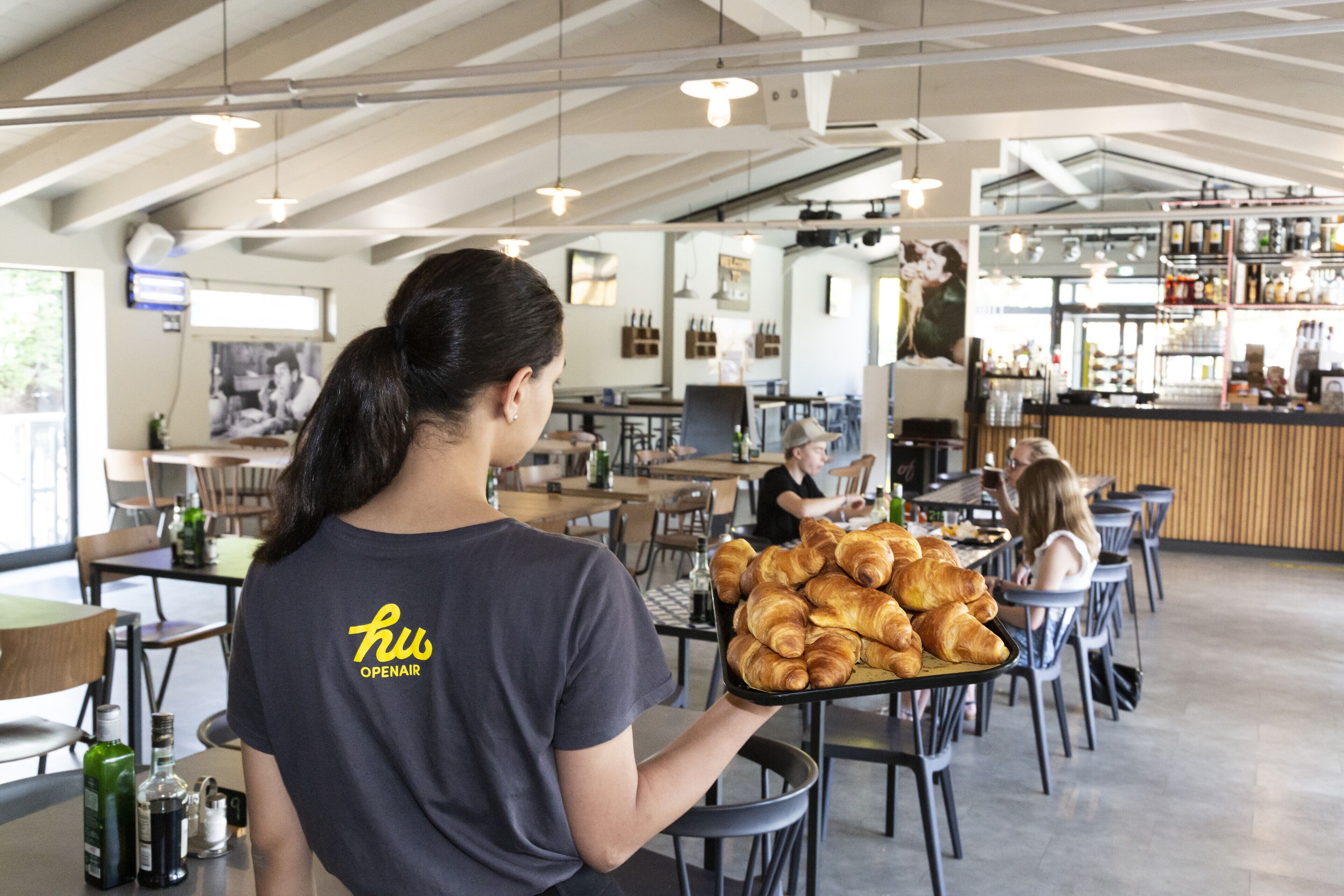 A person holding a tray of croissants in a cafe with tables and chairs, a bar area, and a view of the street outside.