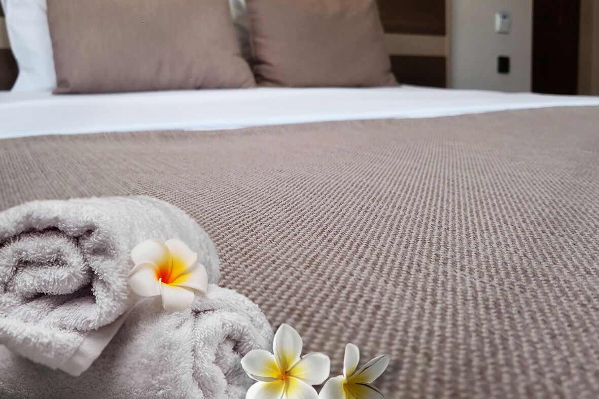 A neatly made bed with a textured beige bedspread, a rolled gray towel, and two white and yellow flowers.