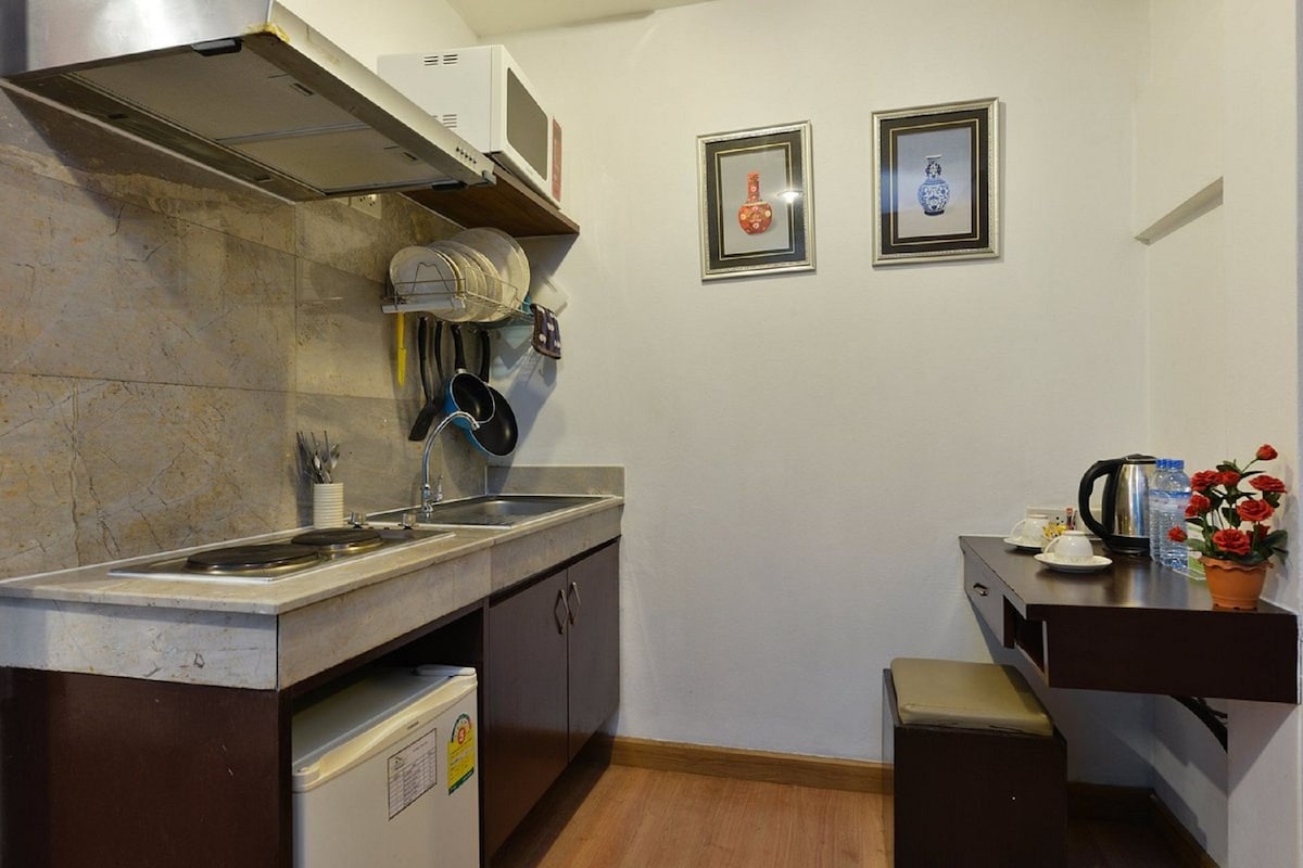 A compact kitchen with a sink, stove, and cabinets. There are two framed pictures on the wall and a small table with a kettle and flowers.