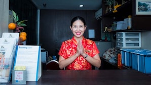A reception desk with a person in a red floral shirt, a sign for booking.com, and a card for taxi service. - Arawana Express Chinatown (Bangkok)