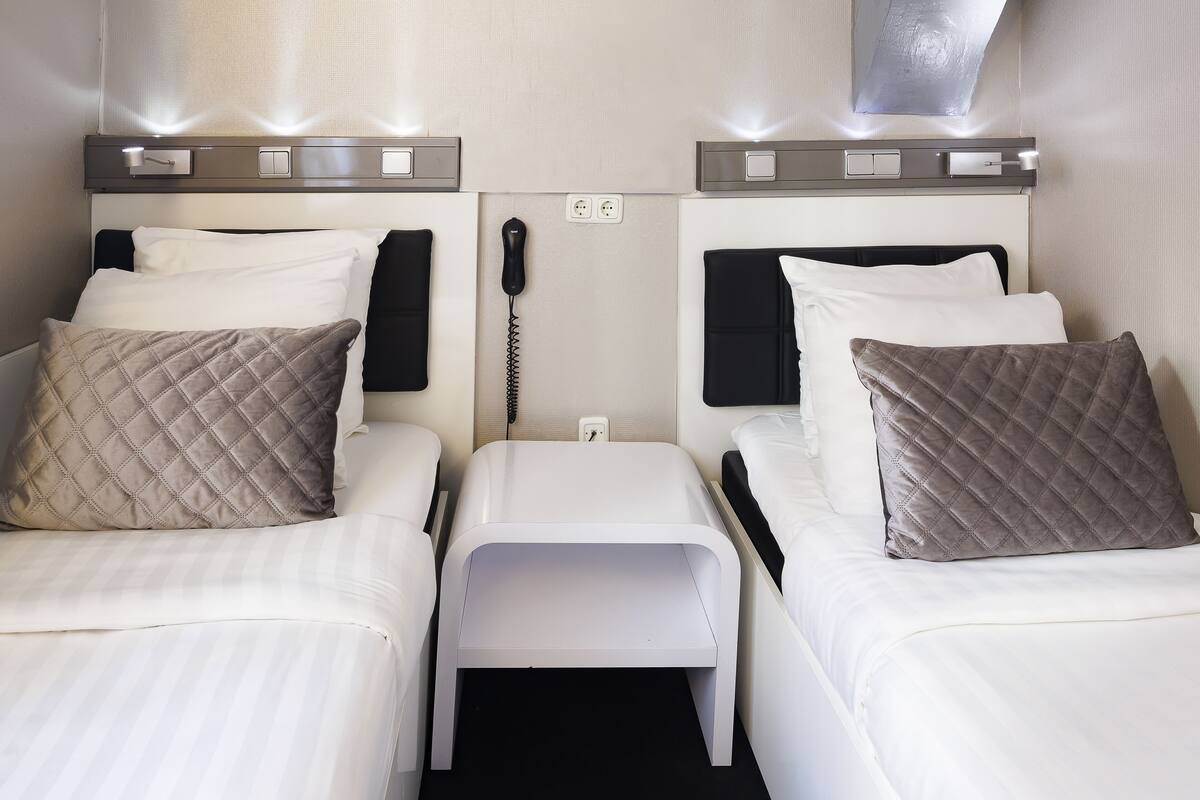A hotel room with two beds, each with a pillow and a bedside table. There is a phone on the wall between the beds.