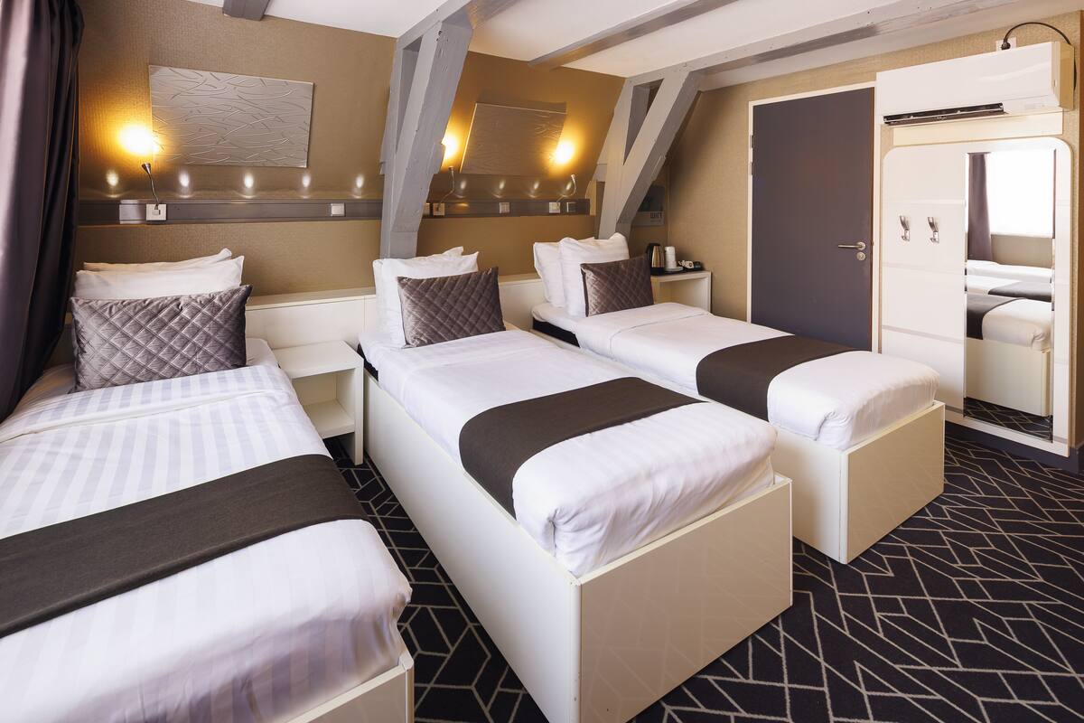 A hotel room with three beds, a wardrobe, a mirror, and a wall-mounted light fixture.
