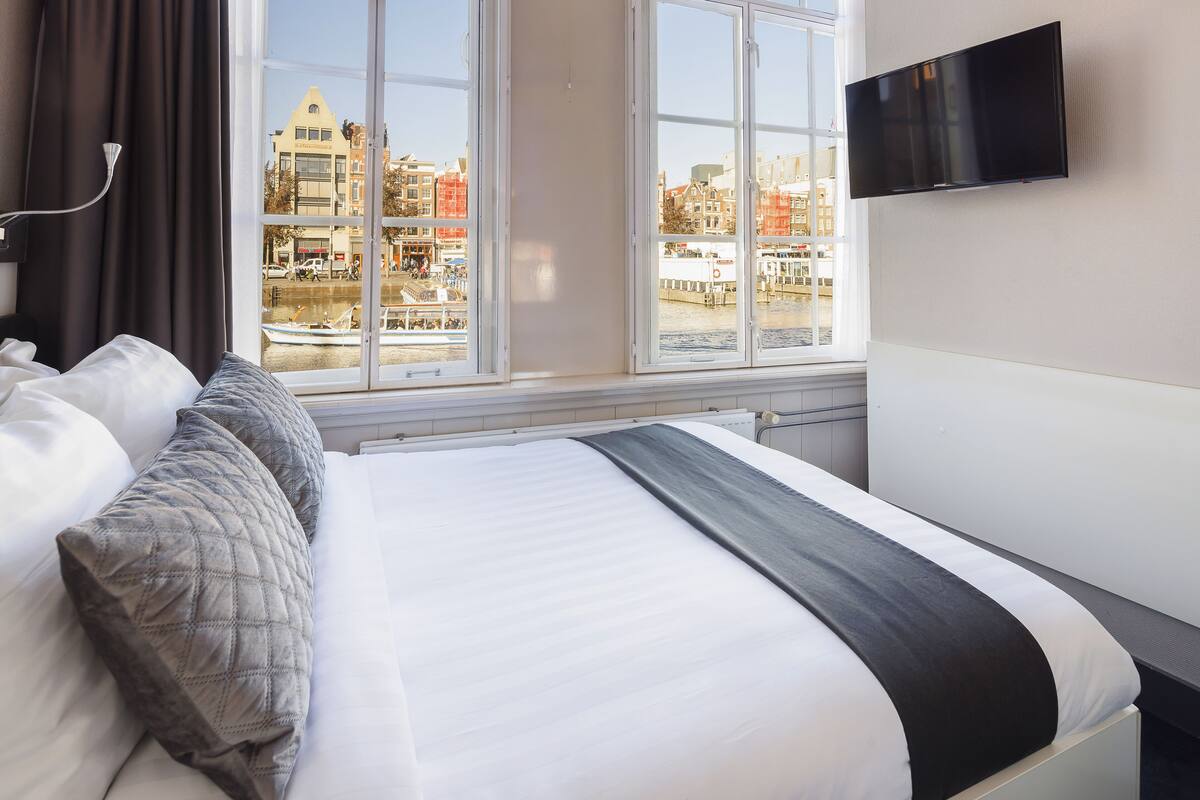 A hotel room with a bed, a TV mounted on the wall, and a view of a cityscape through large windows.