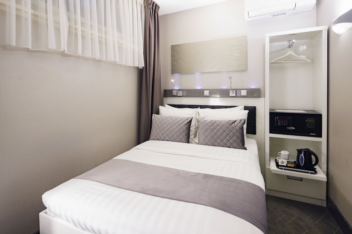 A neatly made bed with a grey and white bedspread, a bedside table with a coffee maker, and a hanging wardrobe.