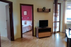 A hotel room with a wooden floor, a bed with white bedding, a TV on a wooden stand, and a window with curtains. - Redenka Holiday Club (Razlog)