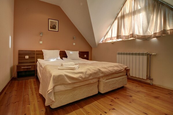 A bedroom with a sloped ceiling, a bed with a headboard, a bedside table, a radiator, and a window with curtains.