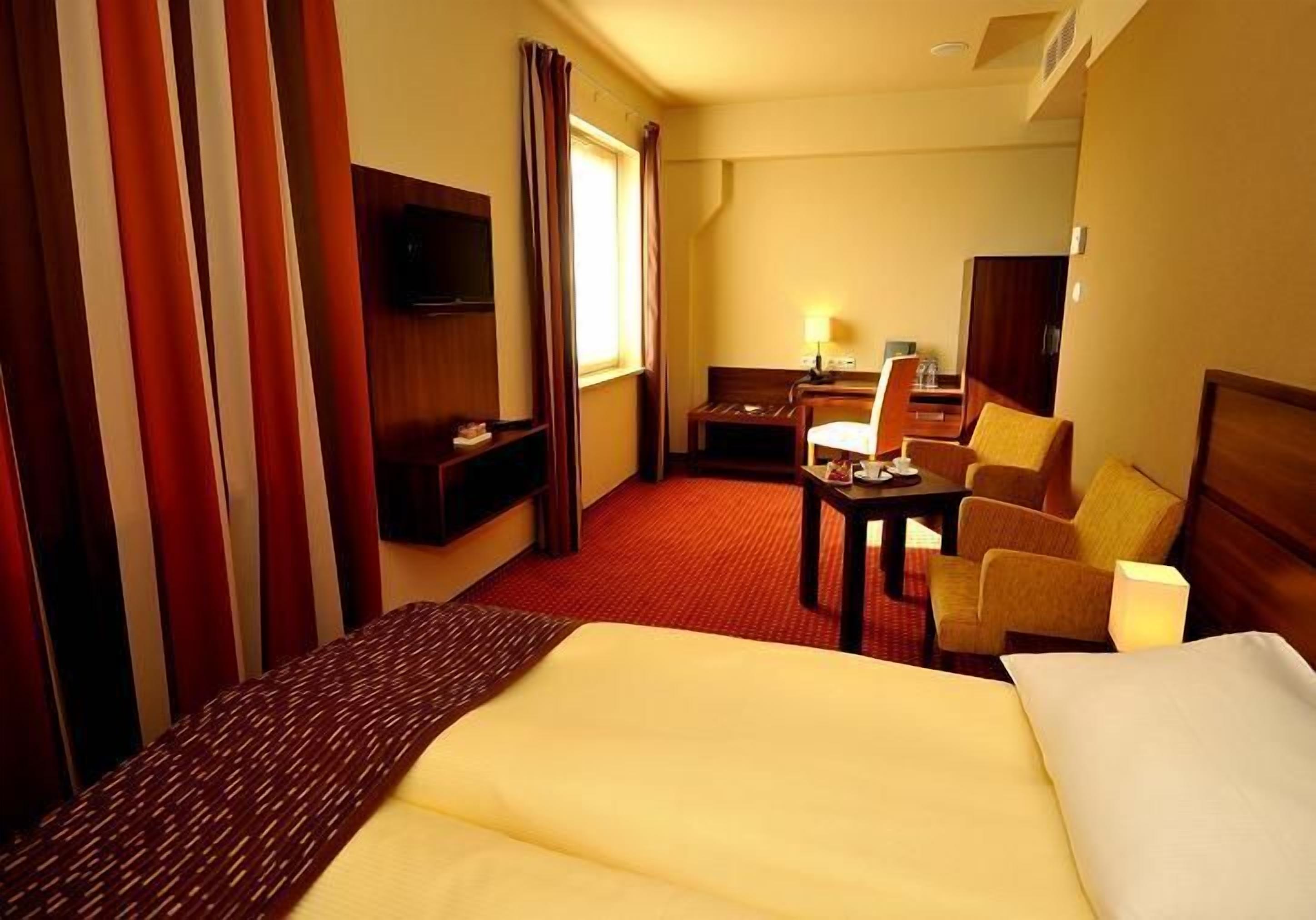 A hotel room with a large bed, a desk with a chair, a TV, and a window with curtains.