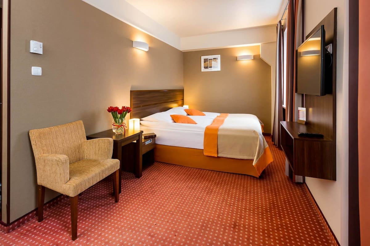 A hotel room with a bed, a chair, a desk, a TV, and a red patterned carpet.