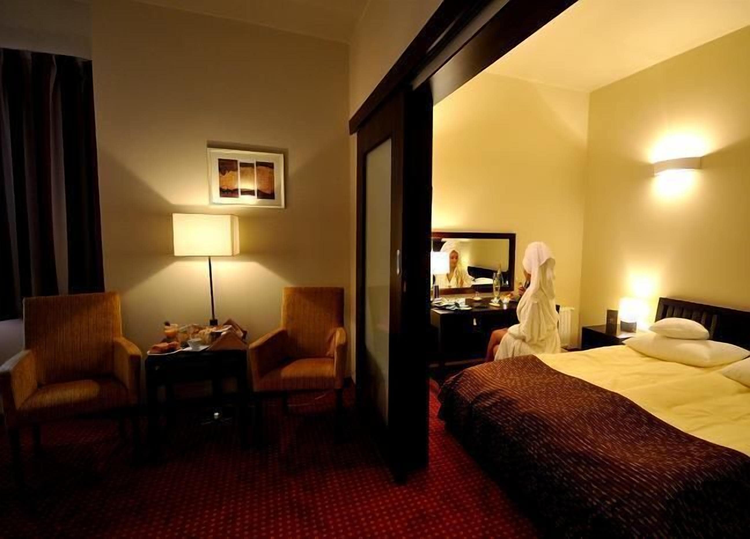 A hotel room with a bed, two armchairs, a desk, a lamp, a mirror, and a picture on the wall.