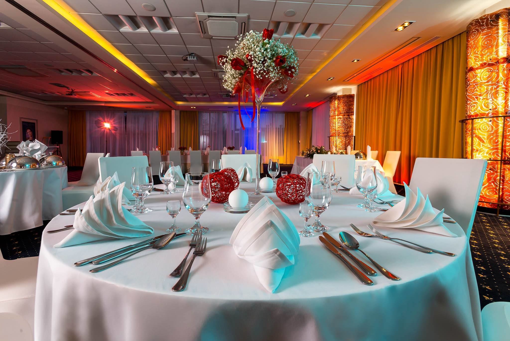 Une salle de banquet avec des tables rondes dressées pour un événement formel, agrémentée de nappes blanches, de serviettes pliées et de couverts élégants.