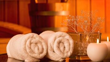 Um espaço de spa com toalhas dobradas, um vaso de vidro com flores secas e velas acesas sobre uma mesa de madeira.