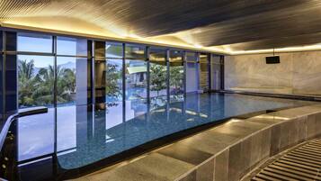 A modern indoor swimming pool with a glass wall, a view of palm trees, and a sleek ceiling design.