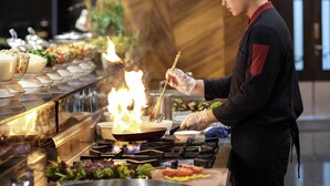 A chef cooking with a pan on a stove, surrounded by various food items and utensils. - Novotel Lampung (Bandar Lampung)
