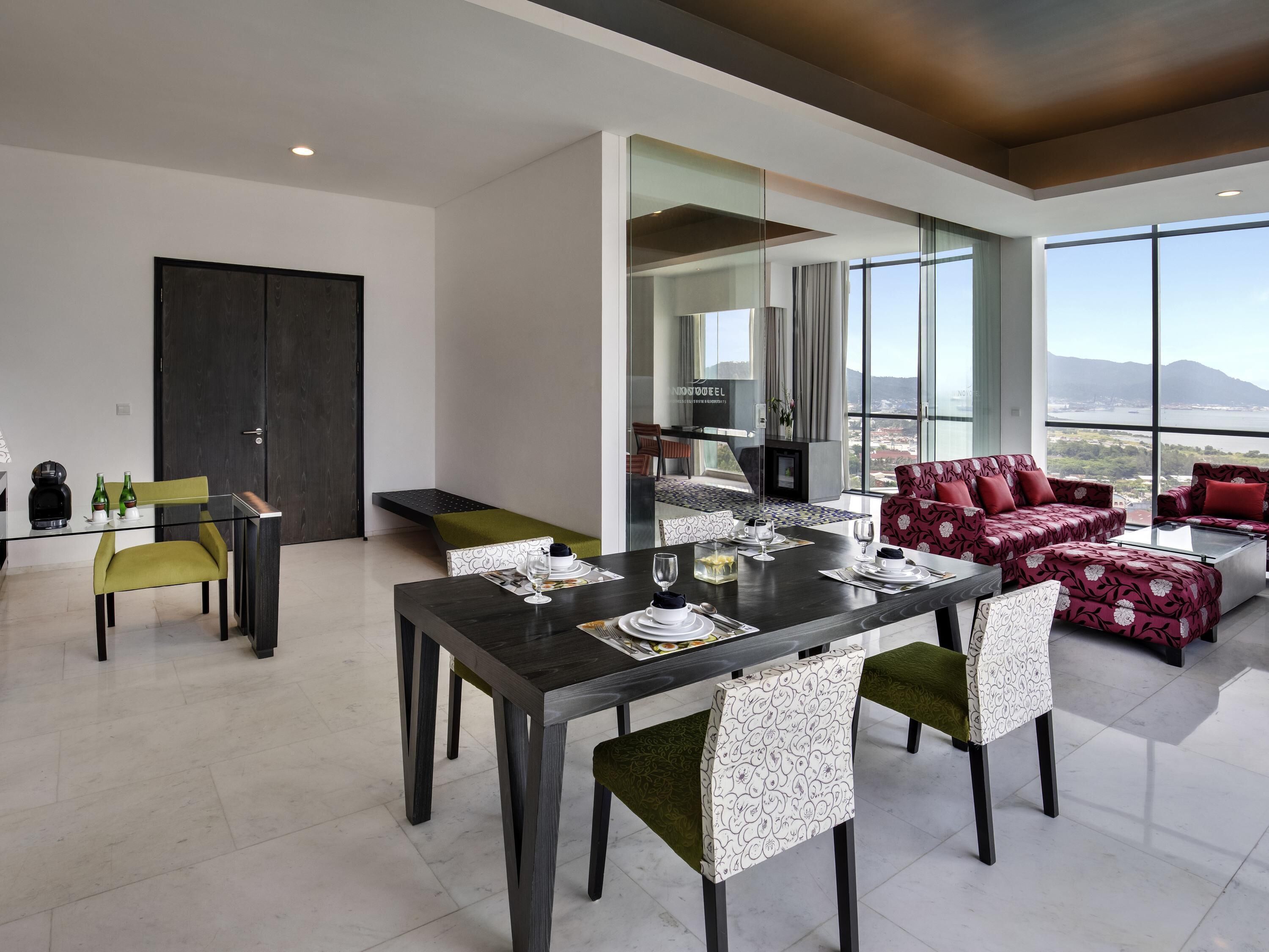 A modern dining area with a dark wooden table, green and patterned chairs, and a seating area with red patterned sofas.