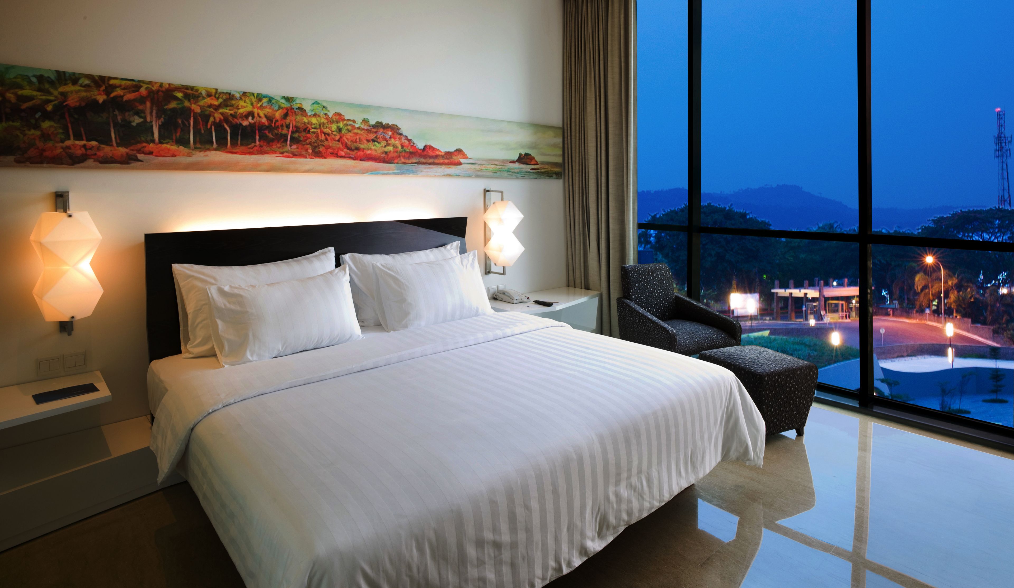 A hotel room with a large bed, two bedside lamps, a nightstand, a chair, and a painting on the wall.
