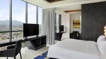 A modern hotel room with a large bed, a TV on a desk, a chair, and a view of a cityscape through large windows.