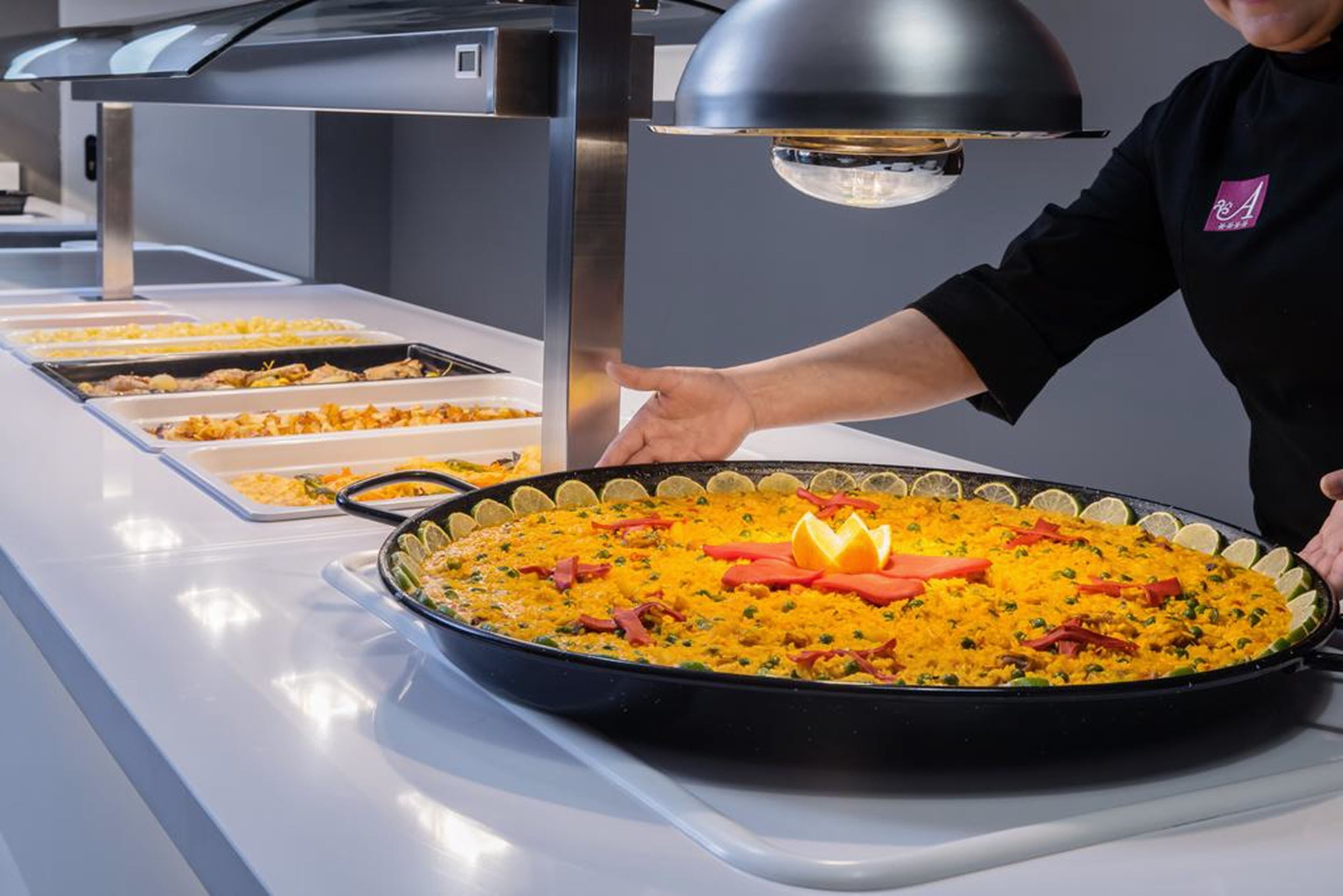 A buffet setup with a large pan of rice and vegetables, a person serving food, and a modern kitchen area with stainless steel appliances.