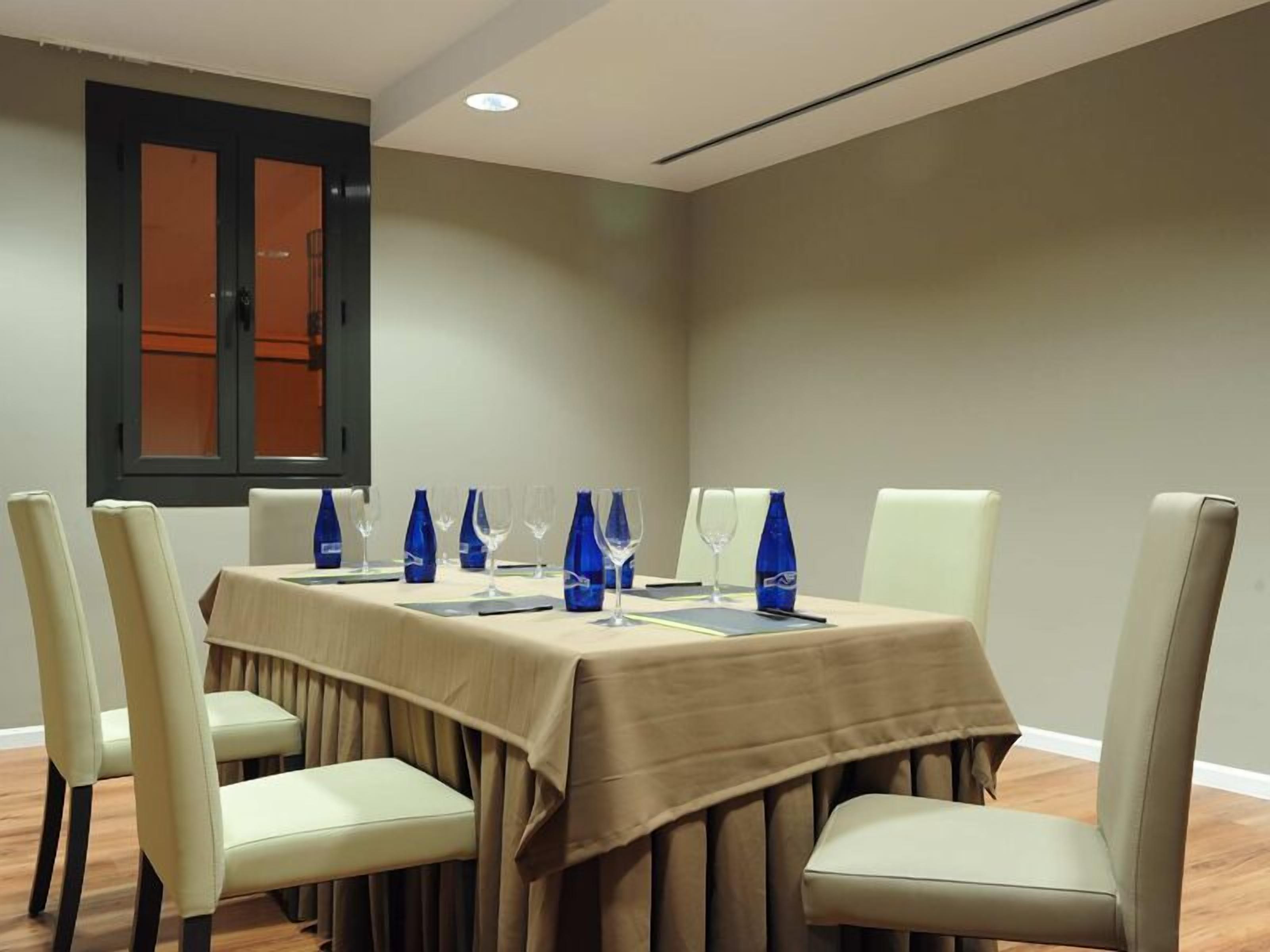 A conference room with a table set for 8, beige chairs, and a window with blinds.