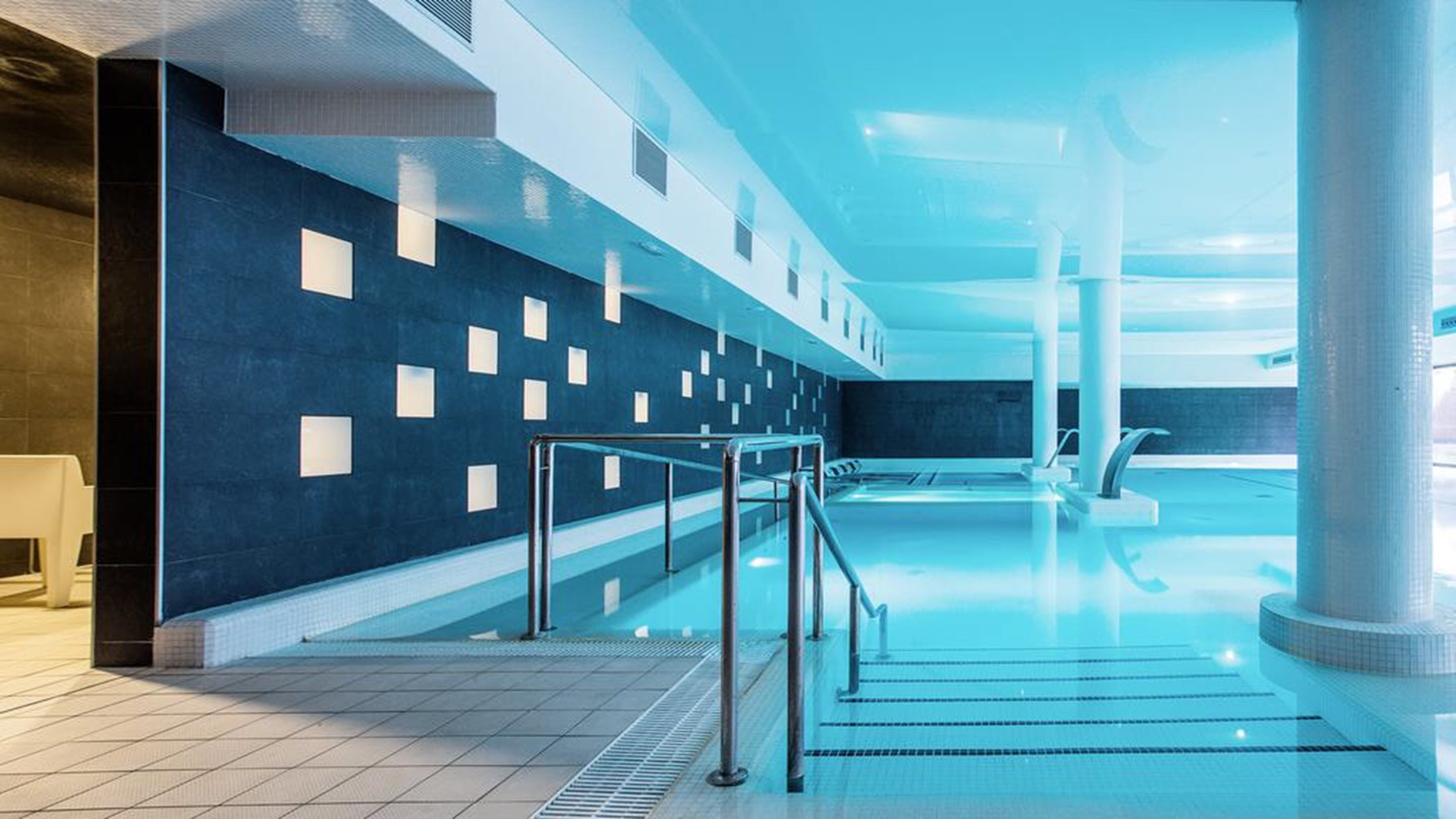 An indoor swimming pool with a modern design, featuring a dark tiled wall with rectangular cutouts and a clear glass barrier.