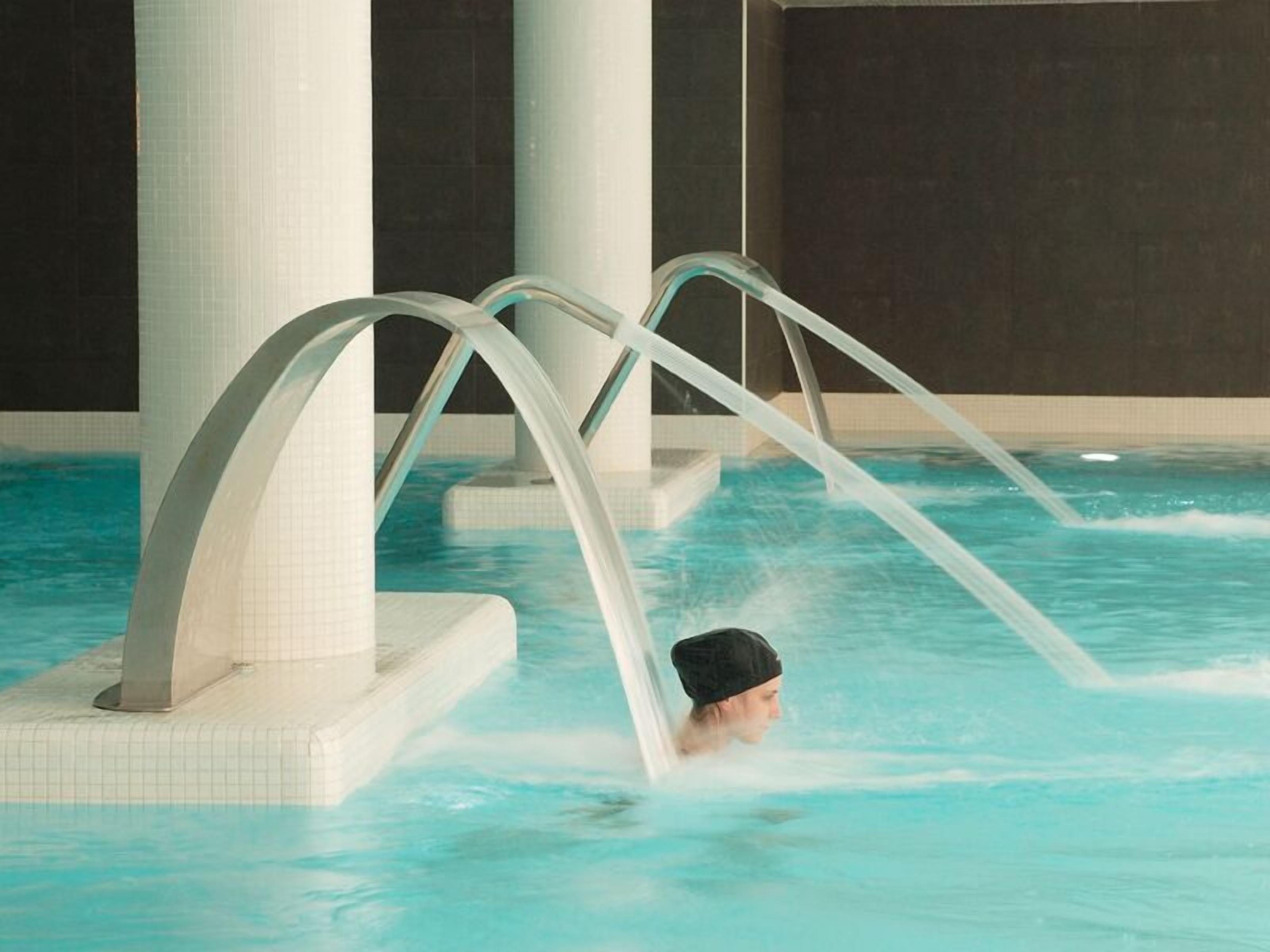 A person in a swimming pool with clear water, surrounded by white tiled walls and columns.