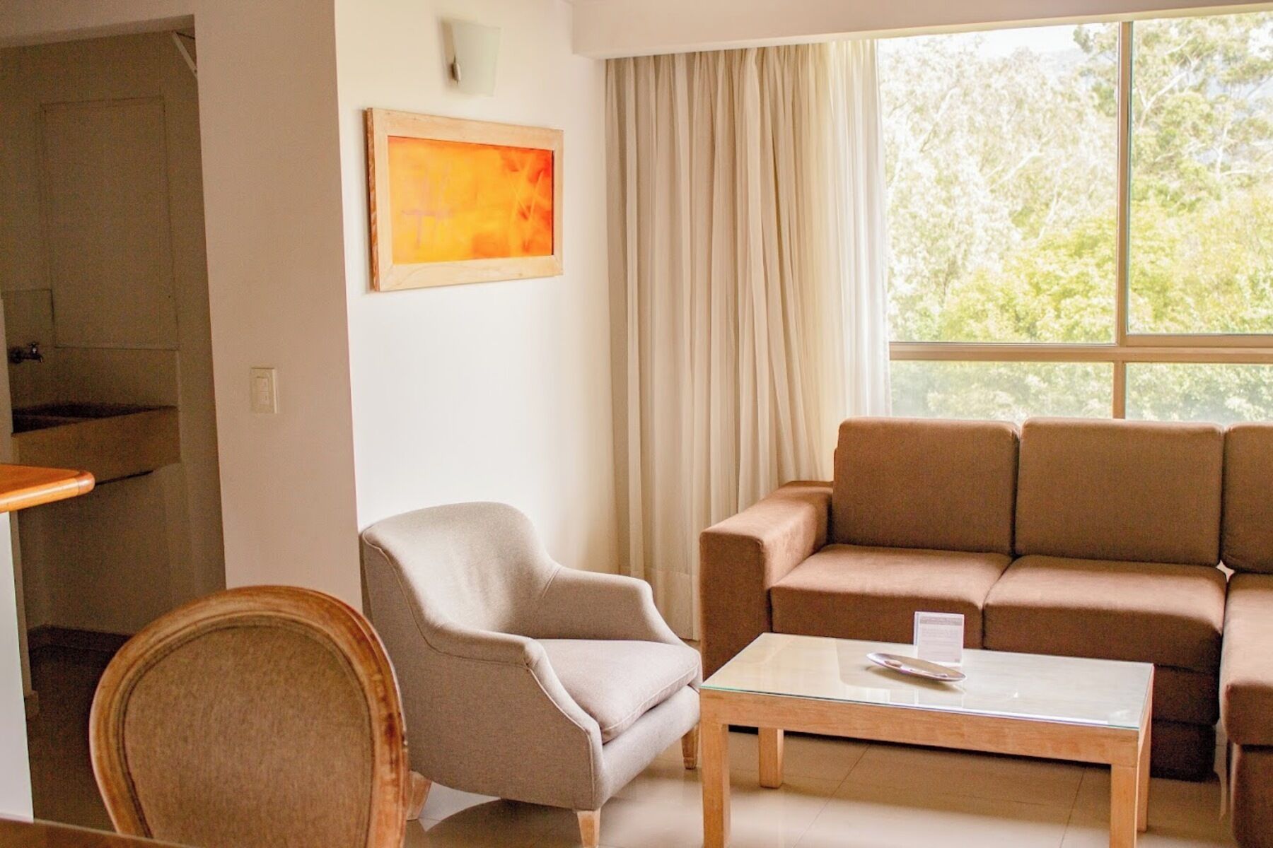 A room with a beige armchair, a brown sofa, and a glass coffee table. There is a painting on the wall and a window with curtains.