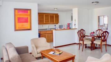 Una sala de estar con sofá, sillones, una mesa de centro y un comedor con mesa y sillas. Se ve una cocina al fondo con armarios de madera y una encimera.