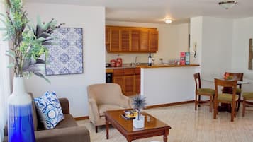 A living room with a sofa, armchair, coffee table, and a dining area with chairs. There is a kitchen in the background with wooden cabinets and a countertop.