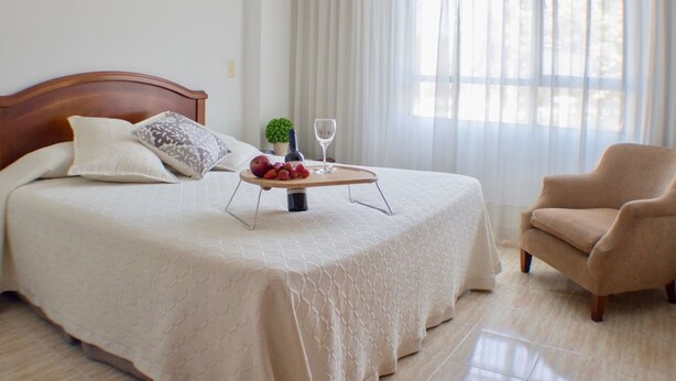 A neatly made bed with a wooden headboard, a bedside table with a bottle and a glass, and a chair in a bright room with a large window.