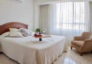 Una cama bien hecha con cabecera de madera, una mesita de noche con una botella y un vaso, y una silla en una habitación luminosa con un ventanal grande.