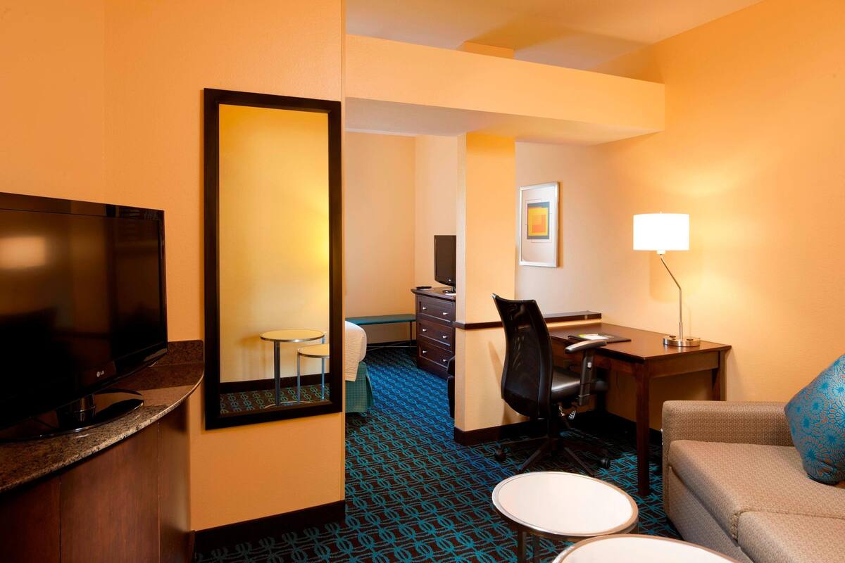 A hotel room with a TV, a desk, a chair, a sofa, a small table, and a mirror.