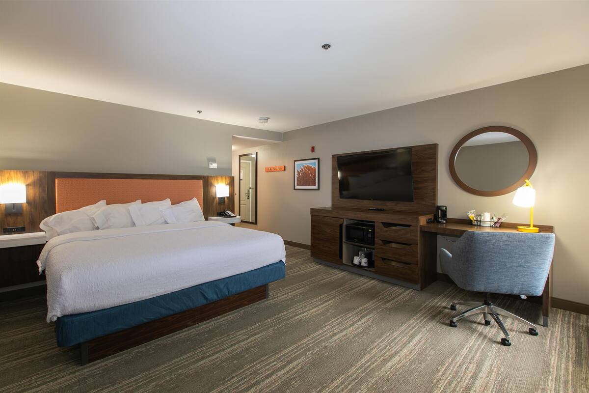 A hotel room with a large bed, a desk with a chair, a television, a mirror, and a lamp.