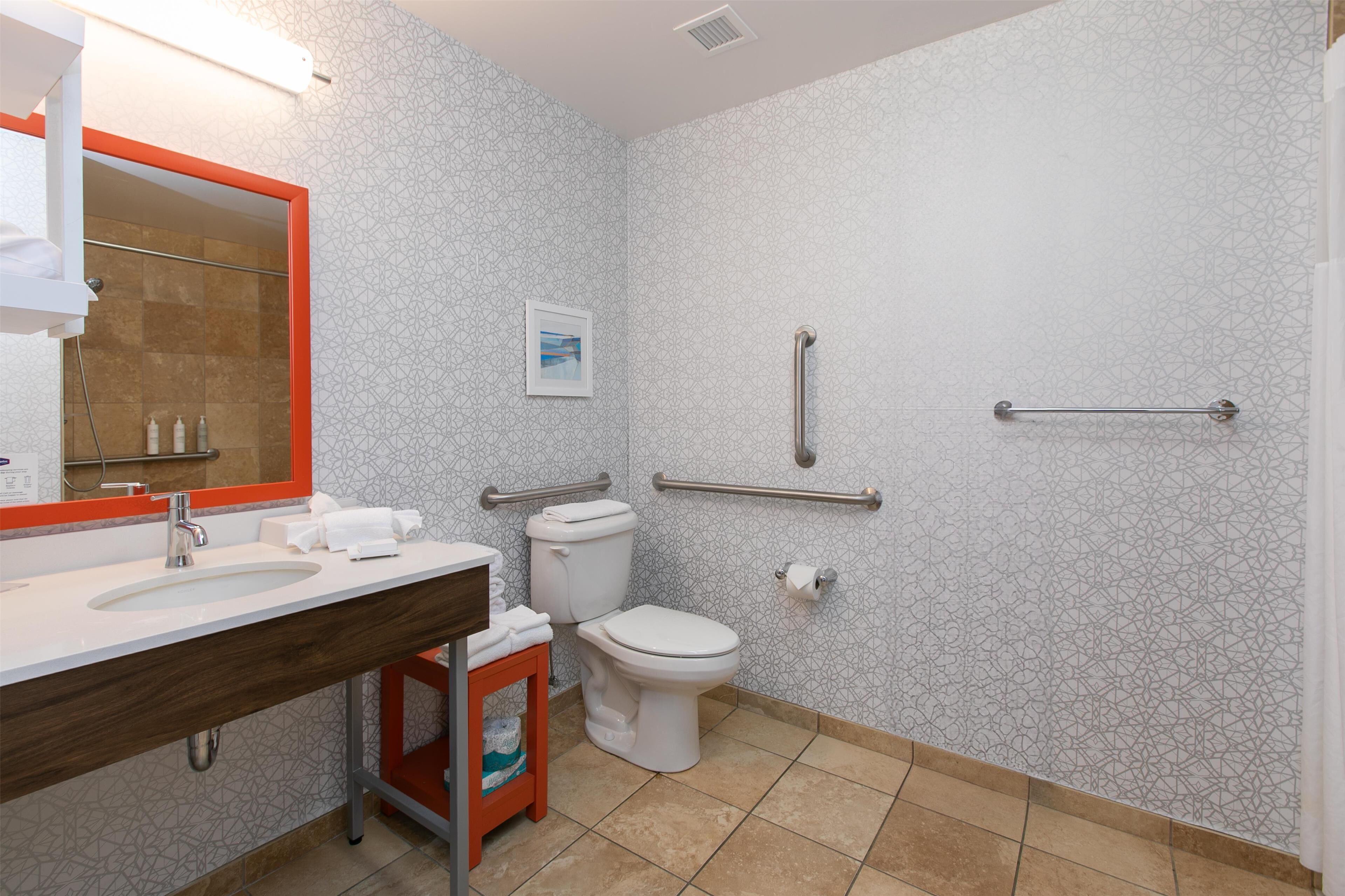 Un bagno moderno con lavandino bianco, water e doccia. Sono presenti mancorrenti alle pareti per sostegno.